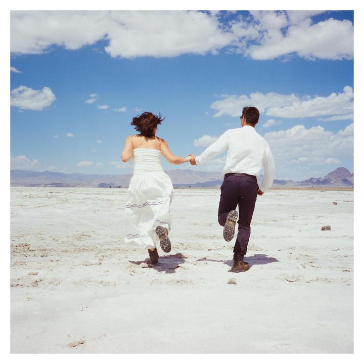 A couple runs hand-in-hand across a vast, arid landscape under a bright blue sky with scattered clouds.