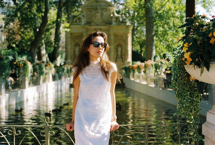 A woman in a white dress and sunglasses stands by a canal with a stone fountain in the background.