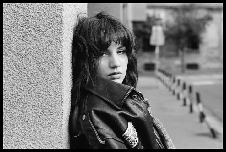 A woman with dark hair and a leather jacket leans against a textured wall and looks at the viewer.