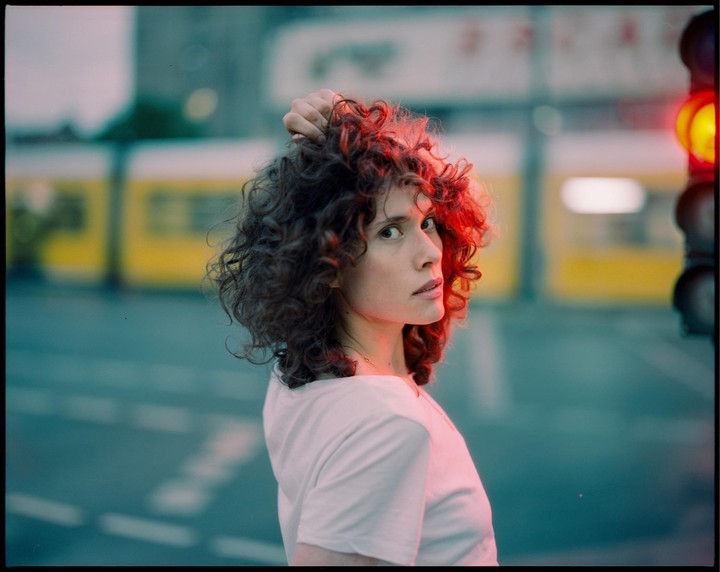 A woman with curly hair looks over her shoulder, illuminated by a red traffic light on a city street.