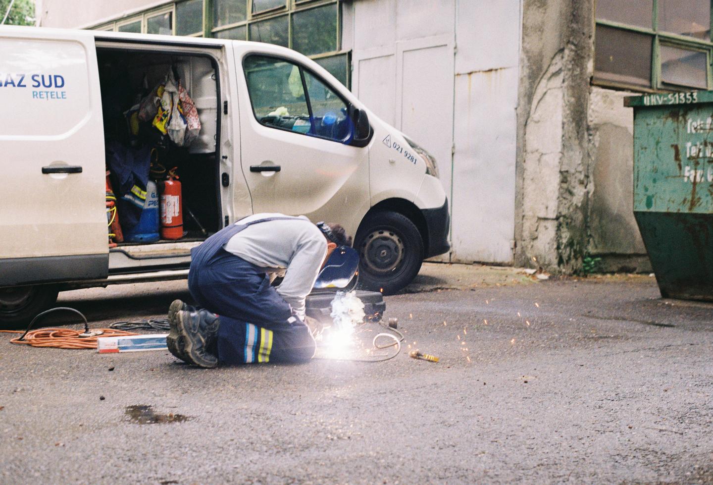 A man in overalls and a welding mask kneels on the ground and welds next to a white van with its side door open.