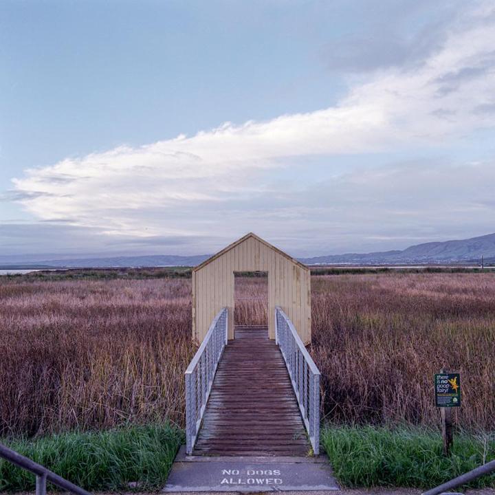 A wooden walkway leads to a small, house-like structure with an open doorway in a field of tall, dry grass under a cloudy sky.