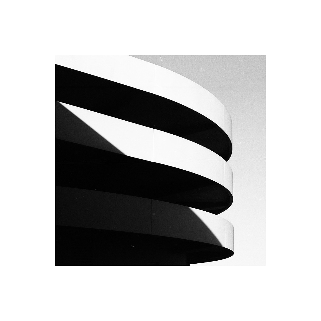 A black and white abstract architectural photograph shows the curved facade of a building with strong shadows.