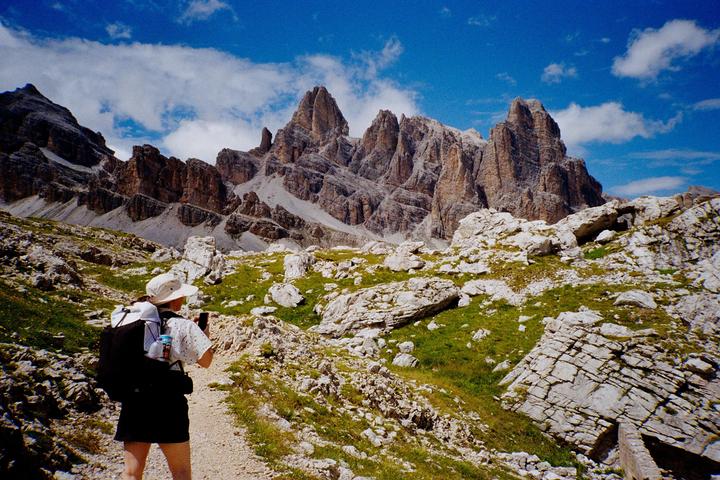 A woman with a backpack and hat stands on a rocky path looking at a mountain range under a blue sky with clouds.