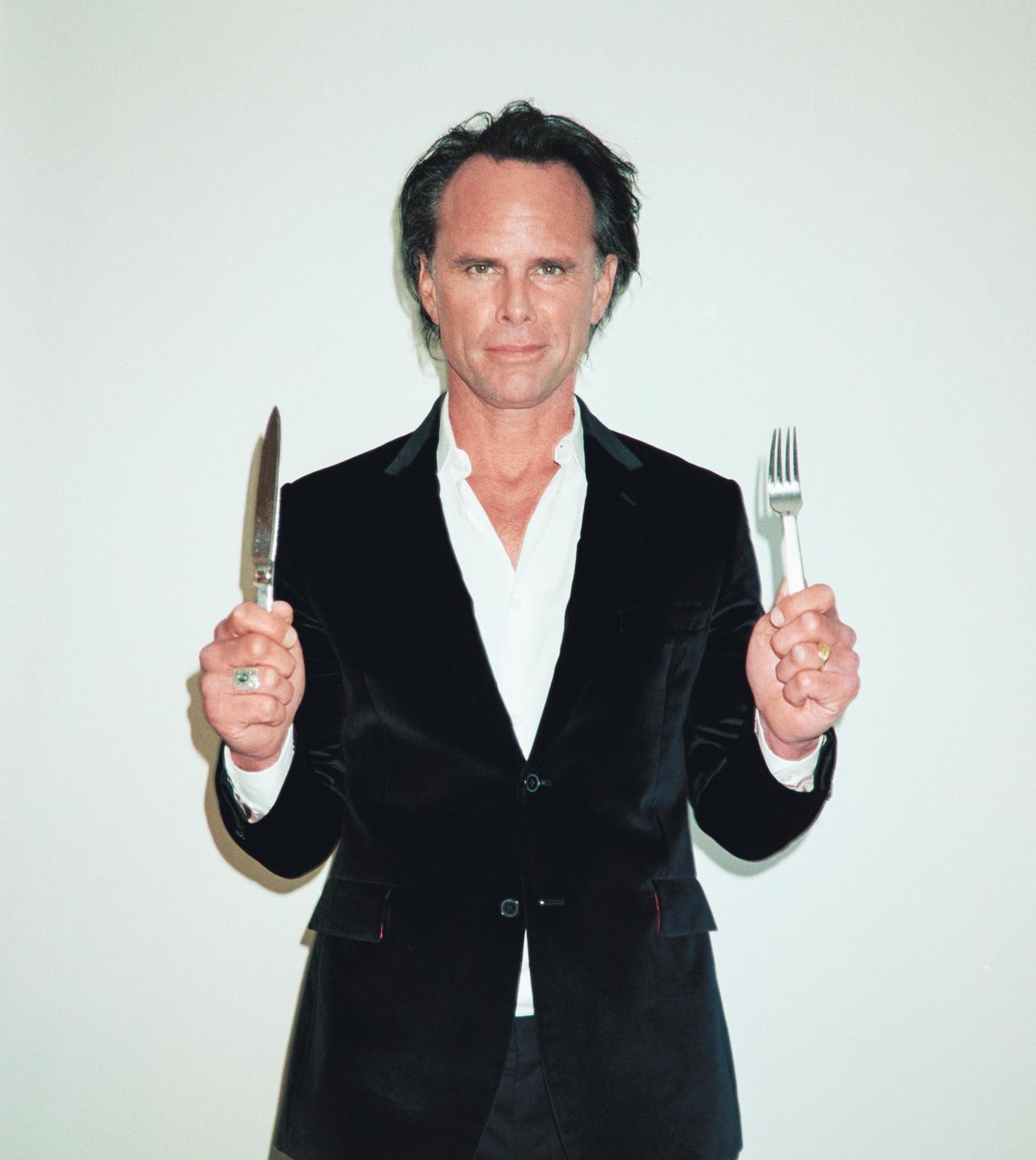 A man in a black velvet jacket holds a knife in one hand and a fork in the other.