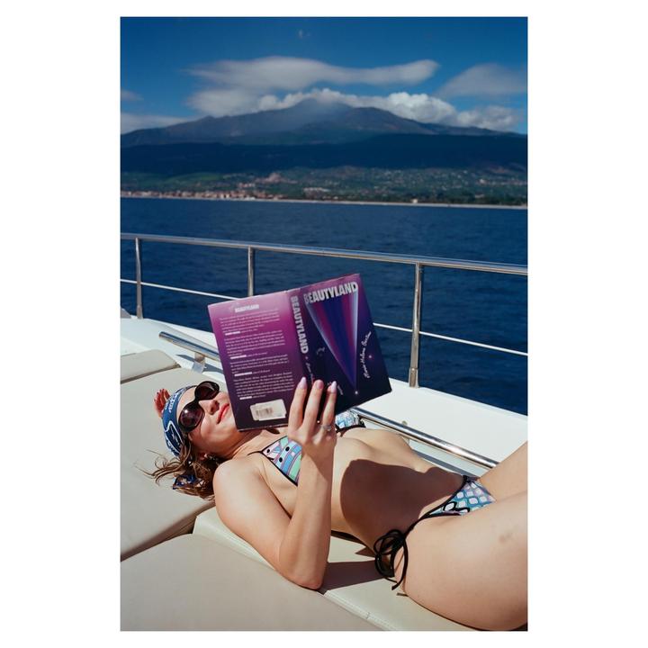 A woman in a bikini and sunglasses reads a book on a boat with a mountain and water in the background.