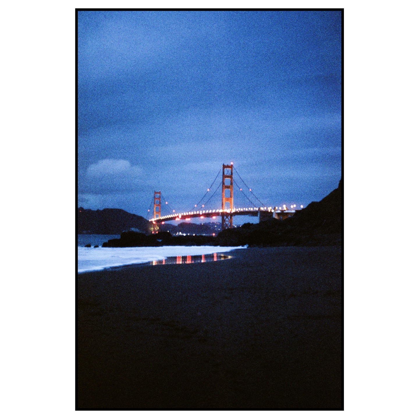 The Golden Gate Bridge is illuminated at night, with its reflection visible on the wet sand of a beach.