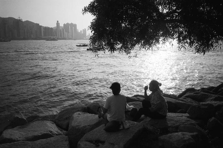 Two people sit on rocks by the water with a city skyline in the distance under a bright sky.