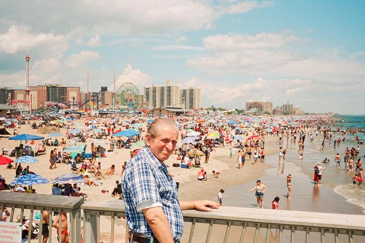 A man in a plaid shirt stands on a pier overlooking a crowded beach with an amusement park in the background.
