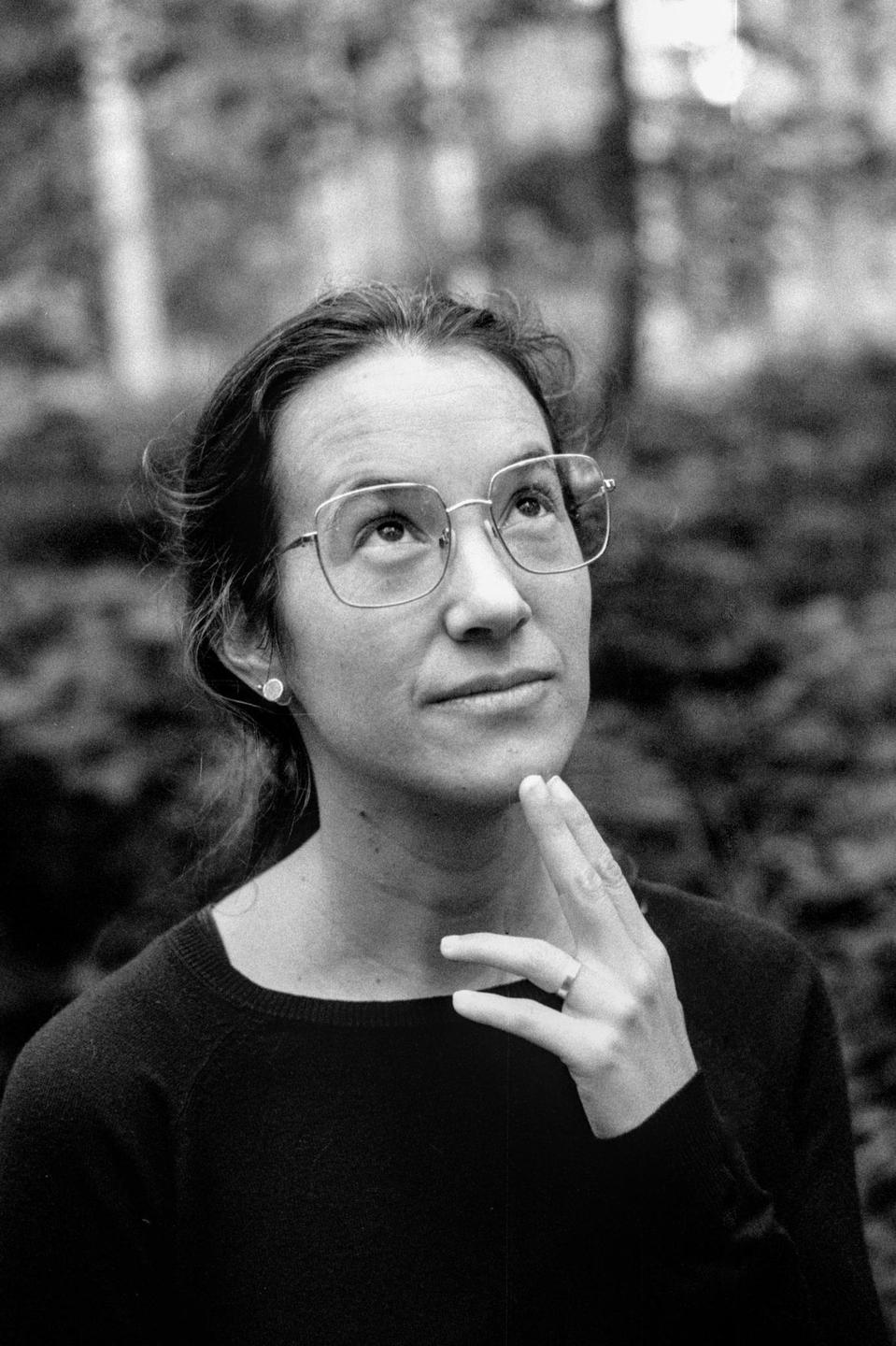 A woman wearing glasses looks up with her hand on her chin in a black and white portrait.