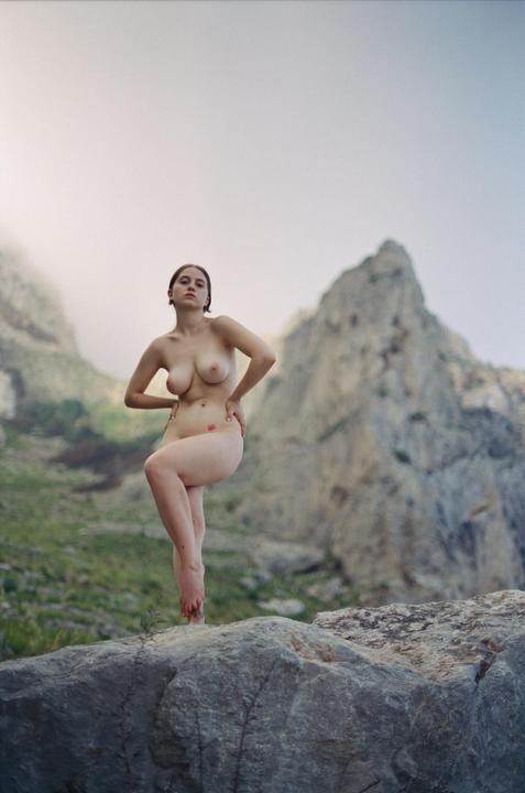 A nude woman stands on a rock with mountains in the background.