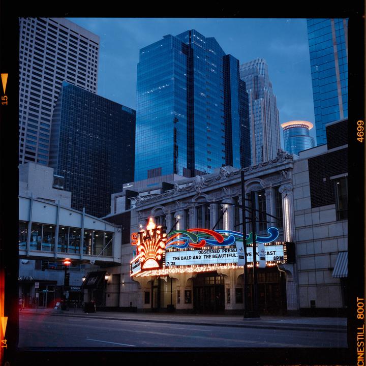 The State Theatre in Minneapolis is illuminated by neon lights at dusk, surrounded by tall buildings.