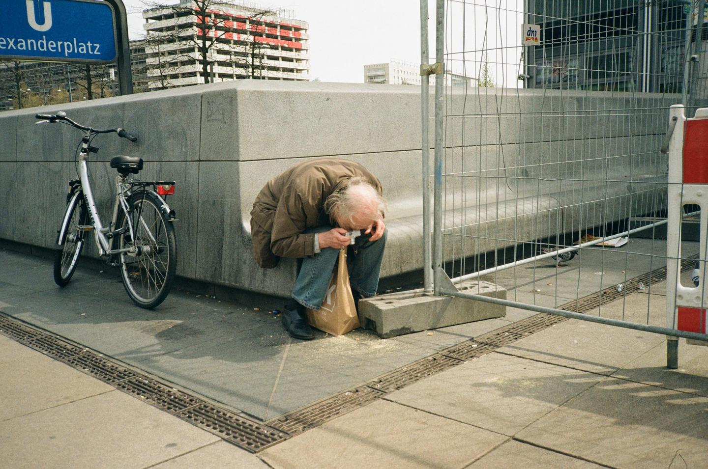 A man bends over a paper bag on the ground next to a bicycle and a construction fence.