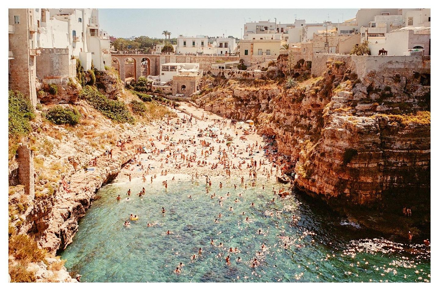 A crowded beach is nestled between rocky cliffs and white buildings under a clear sky.