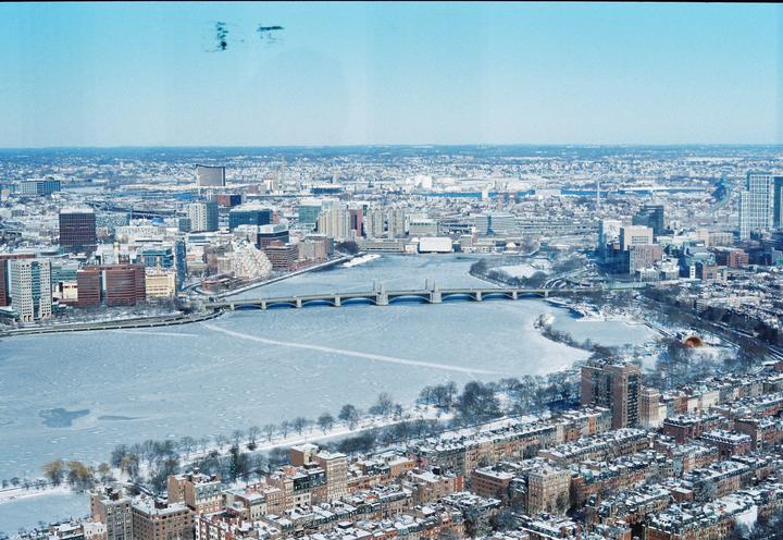 An aerial view shows a frozen river with a bridge and a city covered in snow.