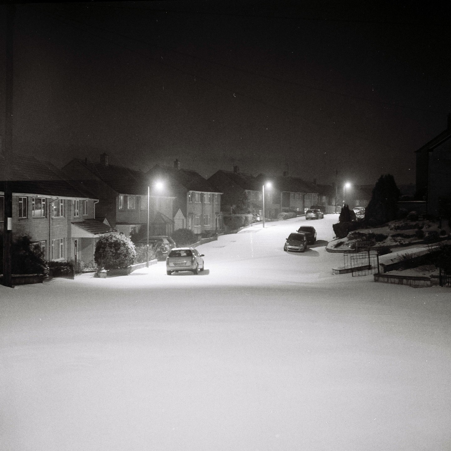 A quiet residential street is covered in snow at night, illuminated by streetlights.