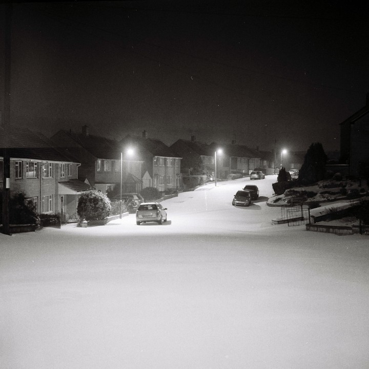 A quiet residential street is covered in snow at night, illuminated by streetlights.