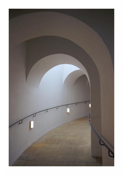 A curved hallway with a handrail and wall lights leads to an arched opening with light coming through.