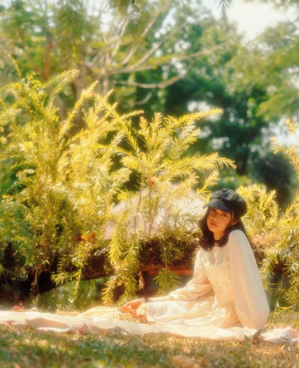A woman wearing a black hat and a white dress sits on a blanket in a garden.