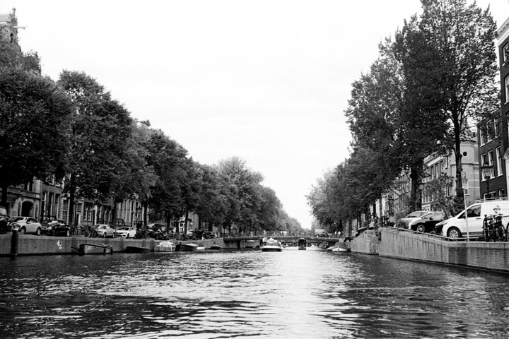 A canal in Amsterdam is lined with trees and buildings, with boats on the water and a bridge in the distance.