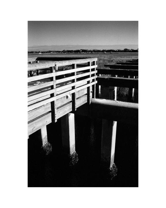 A wooden boardwalk extends over dark water towards a distant grassy landscape under a clear sky.