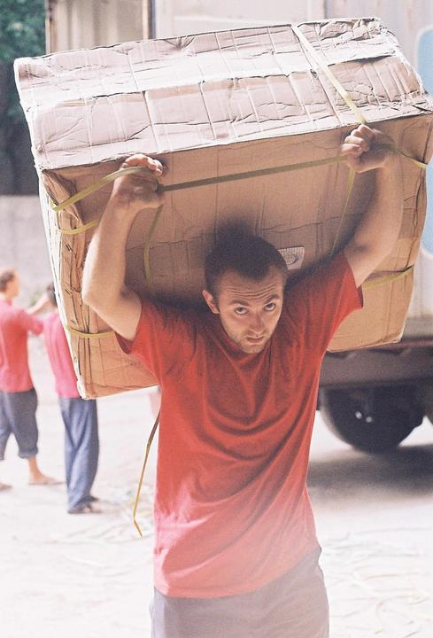 A man in a red shirt carries a large cardboard box on his head, looking directly at the viewer.