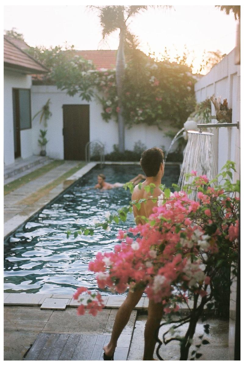 A man showers by a swimming pool with a woman swimming in the background, surrounded by flowering plants.