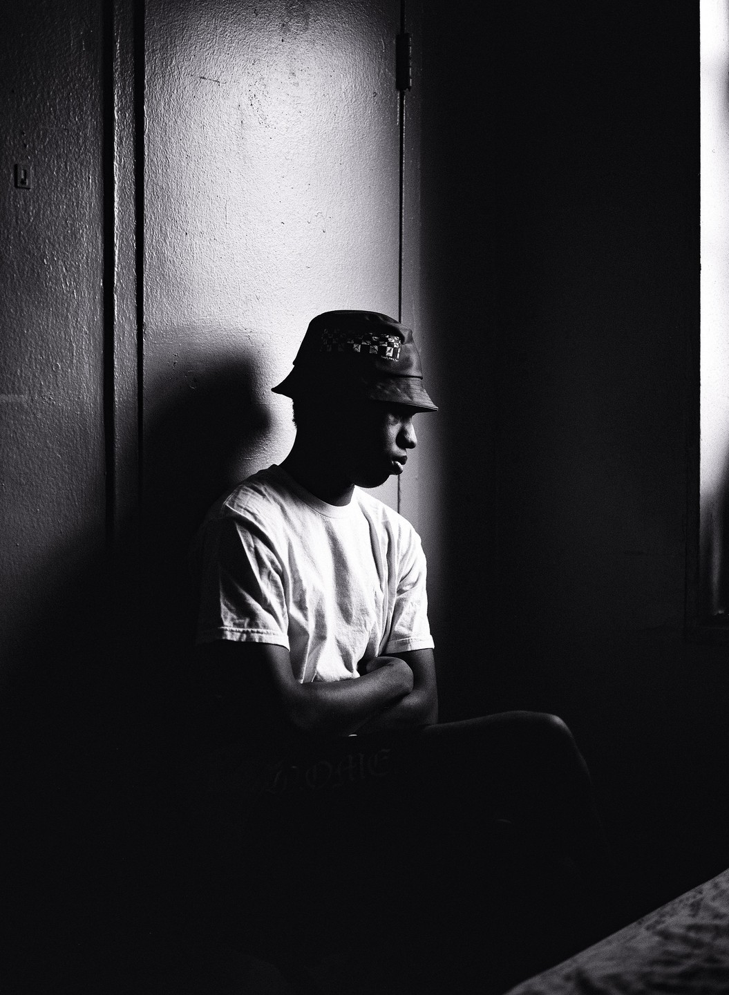A man in a bucket hat and t-shirt sits with his arms crossed in a dimly lit room.