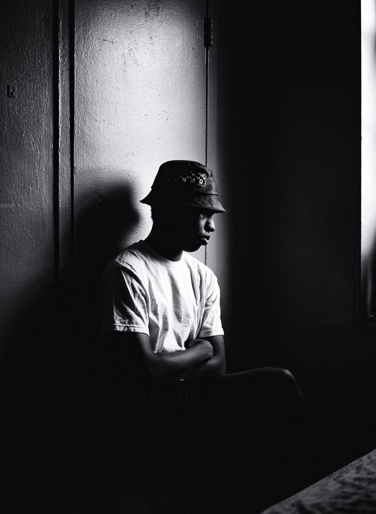 A man in a bucket hat and t-shirt sits with his arms crossed in a dimly lit room.