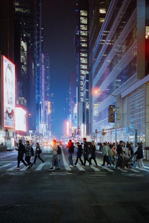 A street in New York City is filled with pedestrians crossing at night, surrounded by tall buildings and bright billboards.