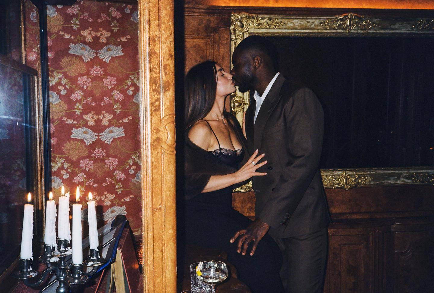A couple kisses in a dimly lit restaurant with patterned wallpaper and a candelabra.