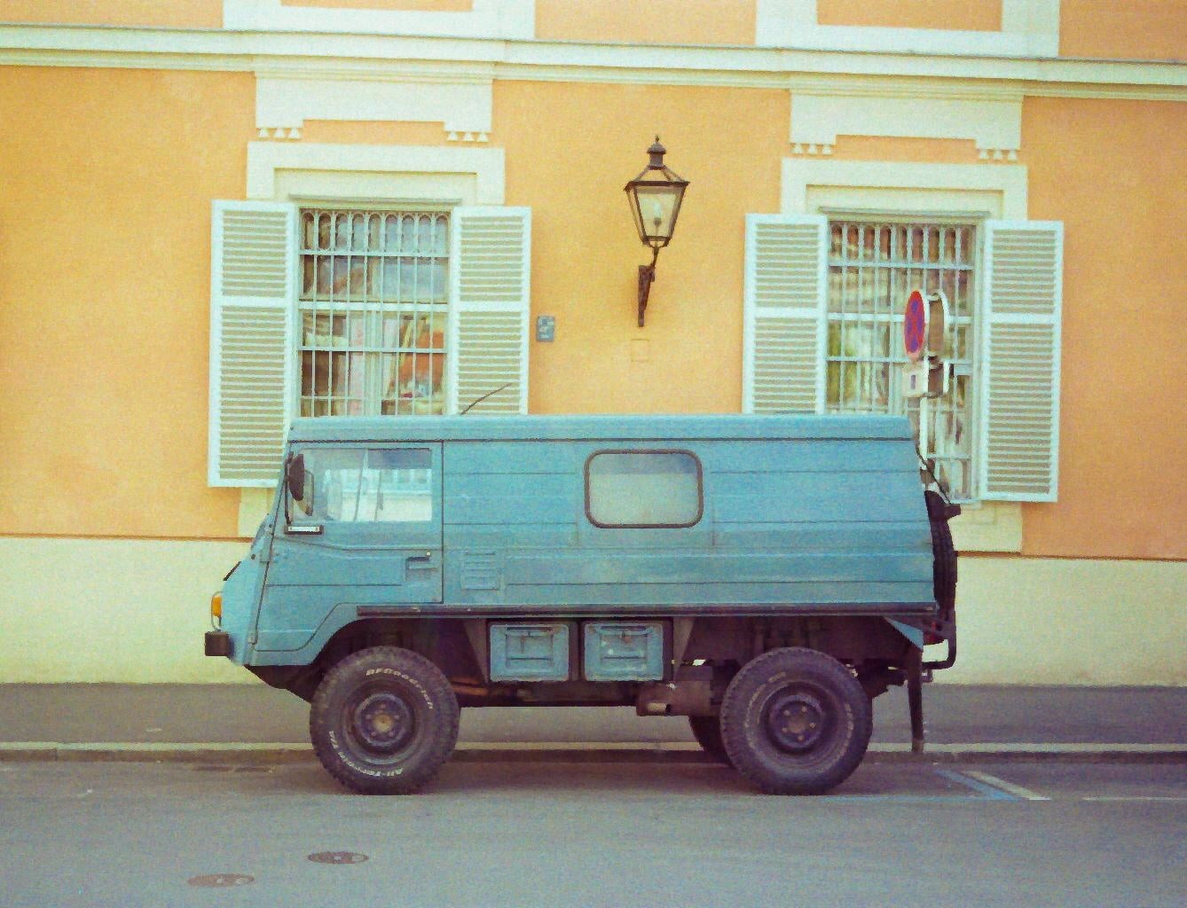 A blue off-road vehicle is parked on a street in front of a building with yellow walls and white window shutters.