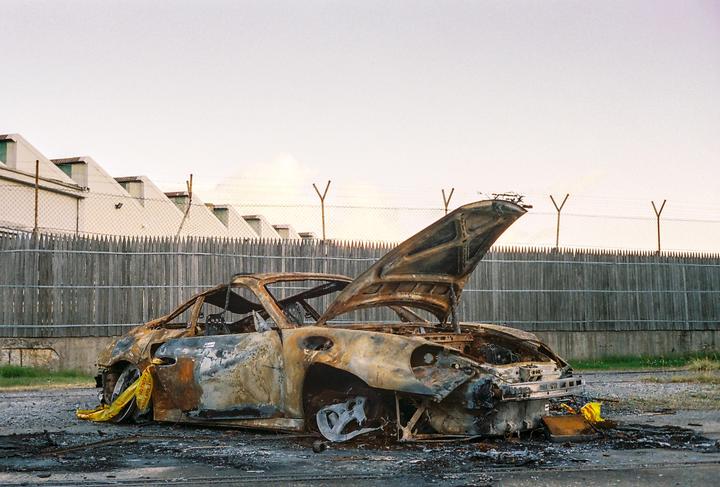 A burnt-out car with its hood open sits on an asphalt lot next to a wooden fence and a building.