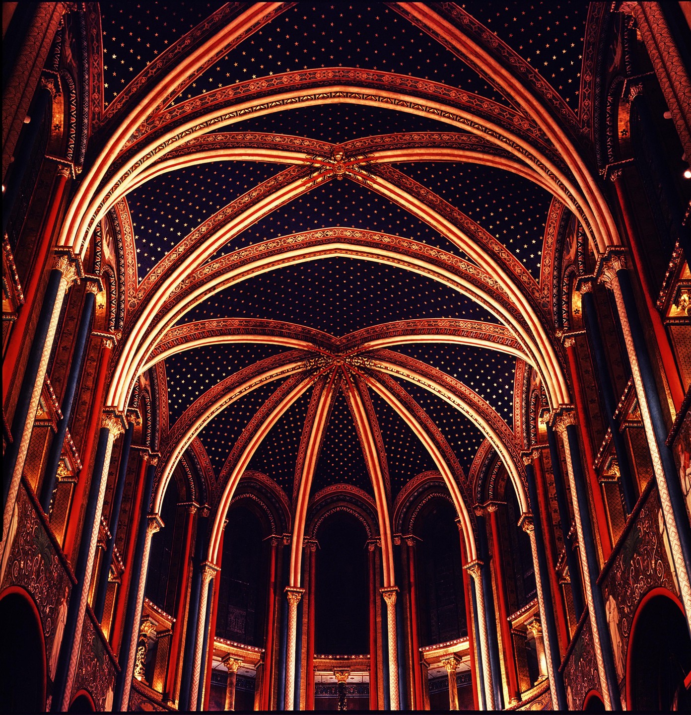 The ornate, vaulted ceiling of a church is illuminated by warm light, revealing intricate patterns and a dark blue background with golden stars.