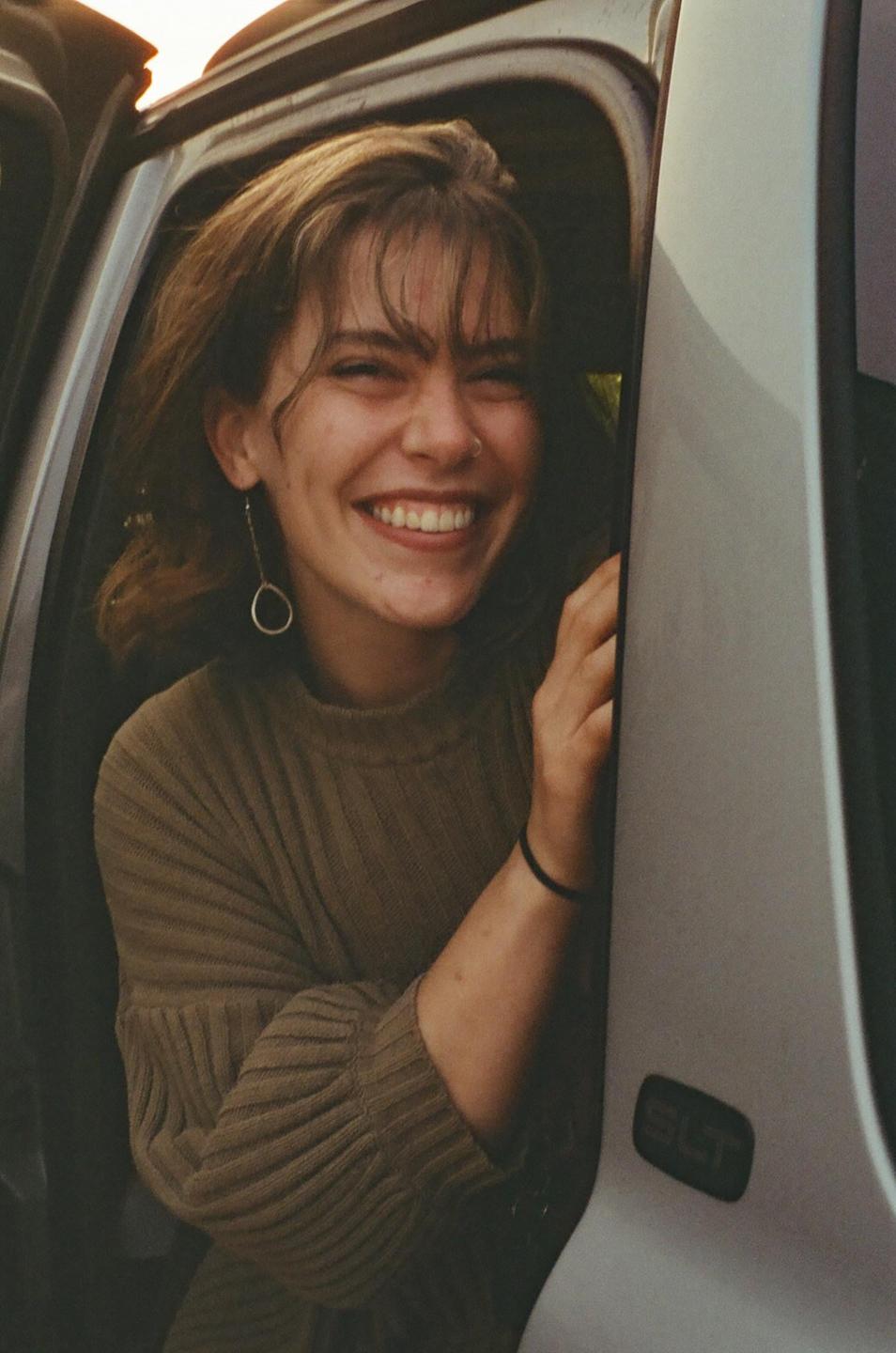 A woman with a nose ring and long earring smiles from the open door of a vehicle.