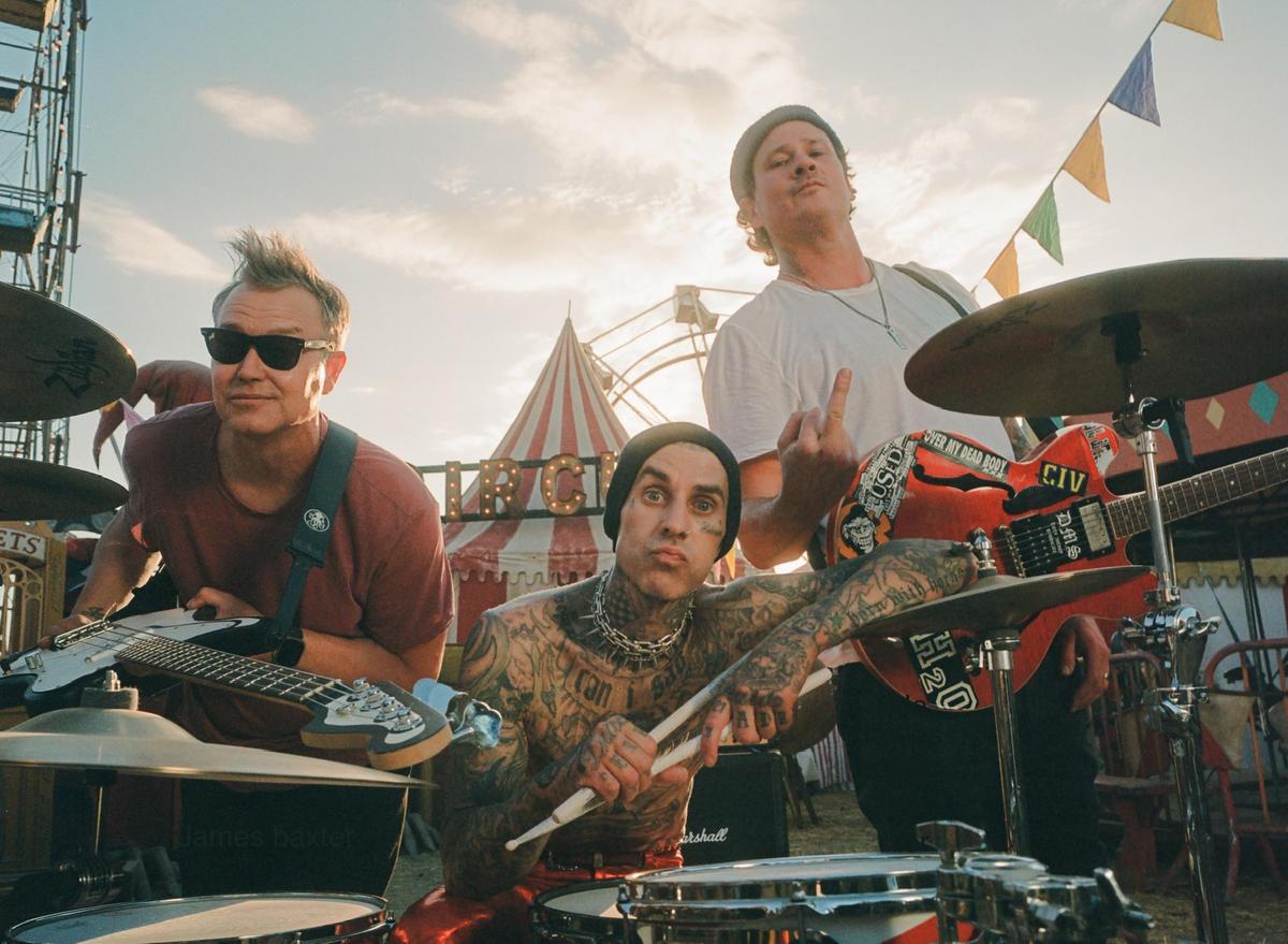 Three men are posing with musical instruments in front of a circus tent and a ferris wheel.