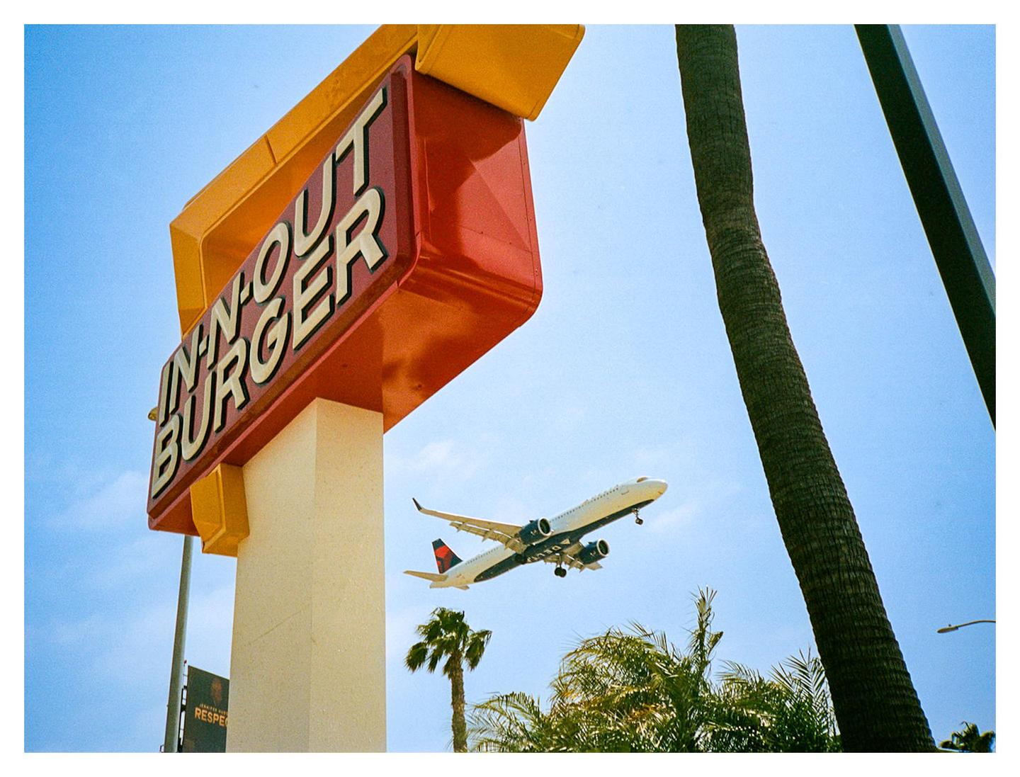 An airplane flies over an In-N-Out Burger sign and palm trees against a clear blue sky.