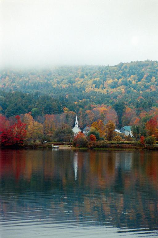 A white church with a tall steeple stands on the shore of a lake, surrounded by autumn trees and reflected in the water.