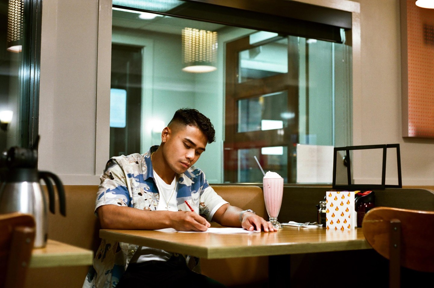 A man sits at a diner booth, writing on a piece of paper next to a milkshake.