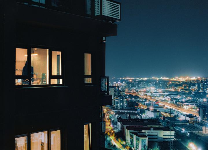 A person is visible through a brightly lit window of a dark building overlooking a city at night.