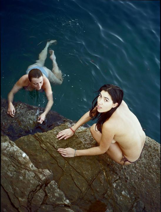 Two women are in the water and on rocks by the sea.