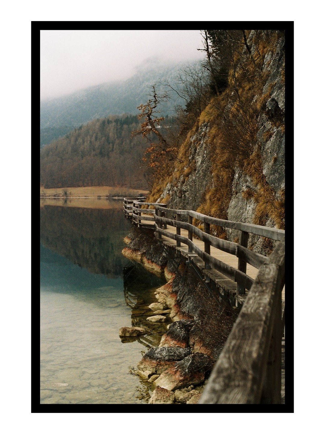 A wooden boardwalk follows a rocky, grassy cliffside next to a lake with a misty mountain in the background.