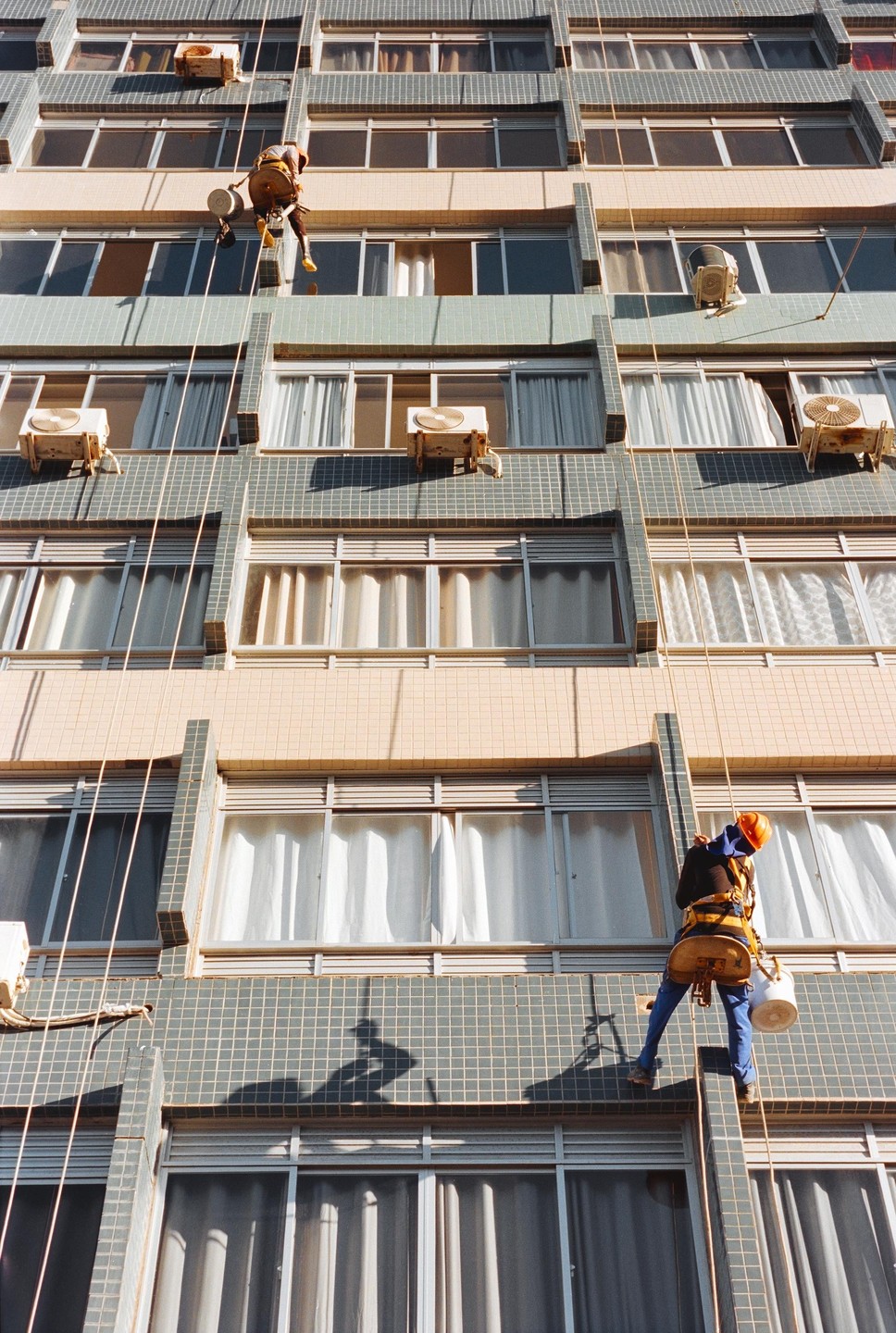 Two men rappel down the side of a tall building with many windows and air conditioning units.
