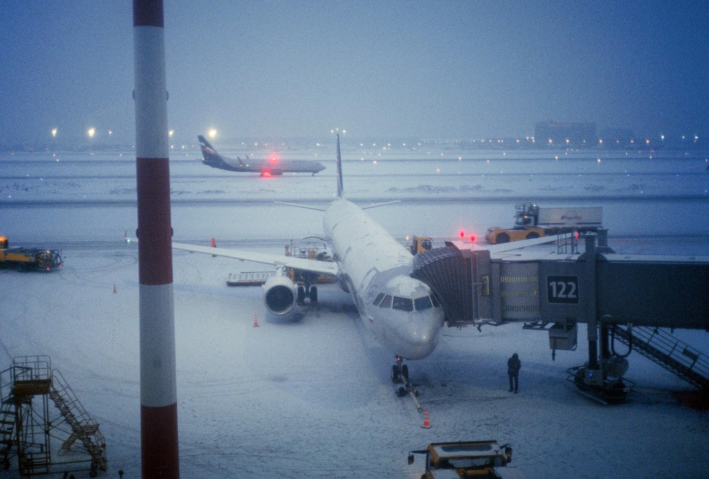 An airplane is parked at a gate on a snowy airfield at night, with another airplane visible in the distance.
