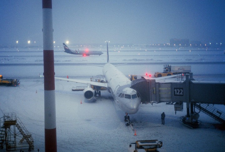 An airplane is parked at a gate on a snowy airfield at night, with another airplane visible in the distance.