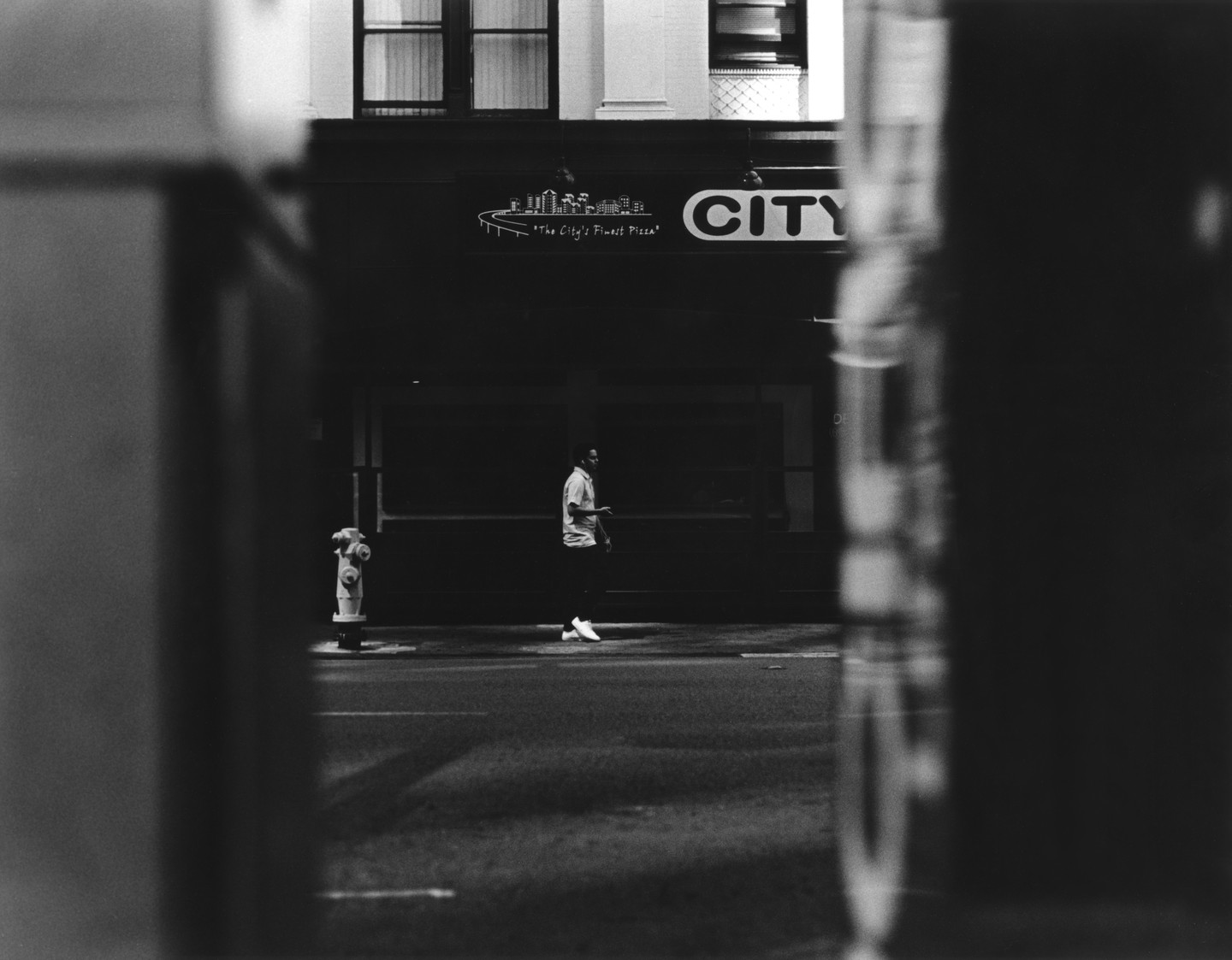 A man walks past a fire hydrant and a pizza shop sign on a city street.