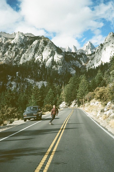 A man on a skateboard rides down a mountain road with a truck parked on the side and pine trees and rocky peaks in the background.