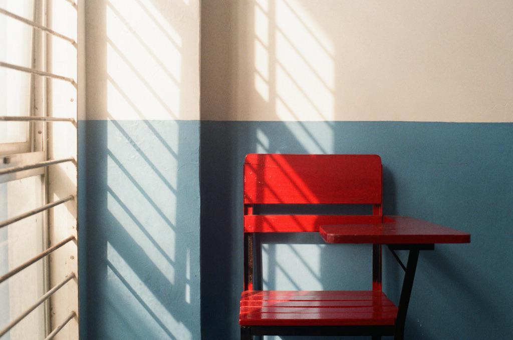 A red school desk sits against a two-toned wall with window shadows cast upon it.