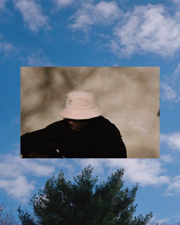 A man in a bucket hat is shown in a rectangular inset against a background of blue sky and green trees.