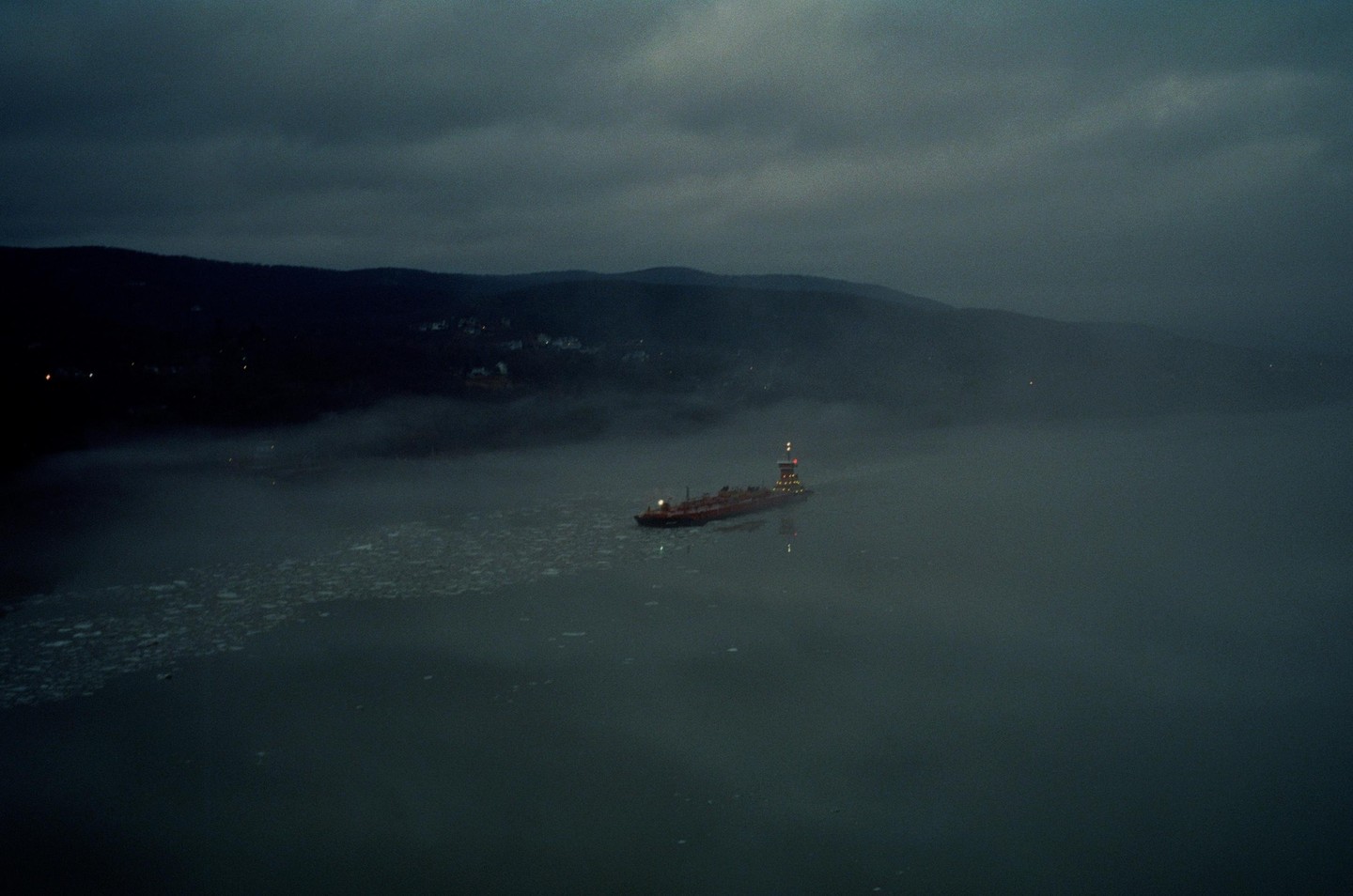 A ship with illuminated lights navigates through a foggy body of water with ice floes, with dark hills in the background.